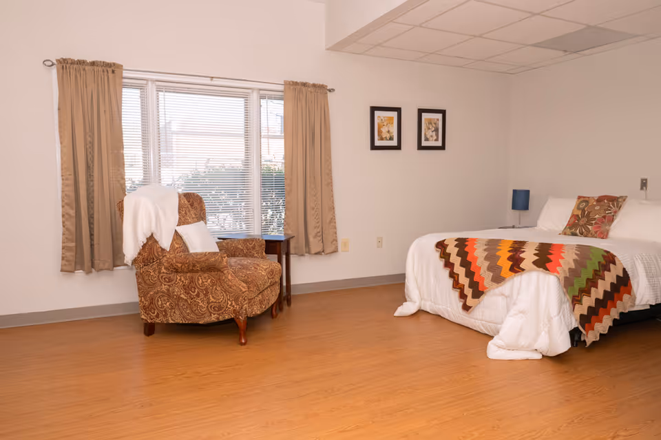 A cozy bedroom with a large window covered by beige curtains. There is a patterned armchair with a white throw draped over it next to a small wooden side table. The bed has white bedding with a colorful zigzag patterned blanket and decorative pillows. Two framed floral pictures hang on the wall above the bed. The floor is wooden and the room has a simple, clean design.