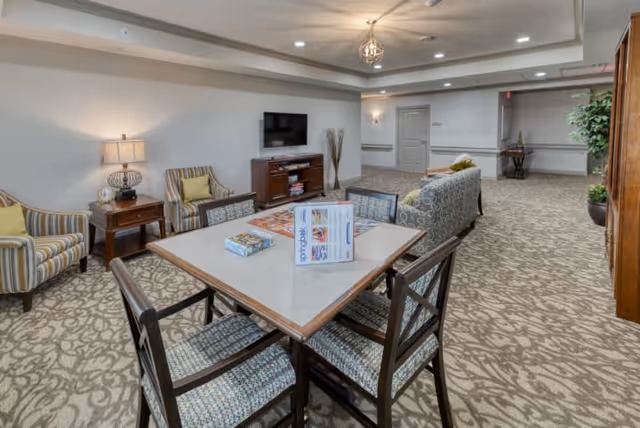 A spacious senior living common area with a square table and four chairs in the foreground. On the table are some brochures and a board game. In the background, there is a TV mounted on the wall above a wooden cabinet, two striped armchairs with yellow cushions, a side table with a lamp, and a patterned sofa. The room has beige patterned carpet, neutral walls, recessed lighting, and decorative ceiling molding.