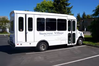 A white Suncrest Village Retirement Community shuttle bus parked in a sunny parking lot.