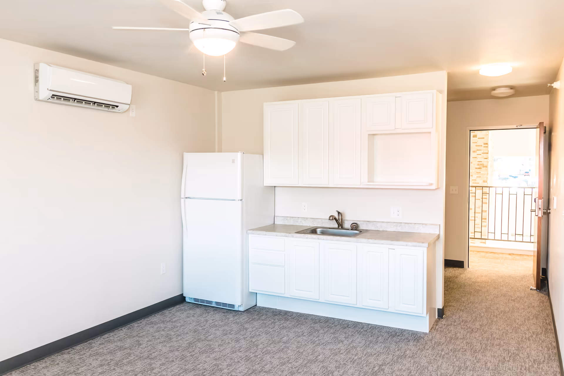 A small kitchen area in an assisted living facility with white cabinets, a white refrigerator, a countertop with a sink, and a ceiling fan with a light. The room has beige walls and carpeted flooring, with an open door leading to an outdoor balcony.