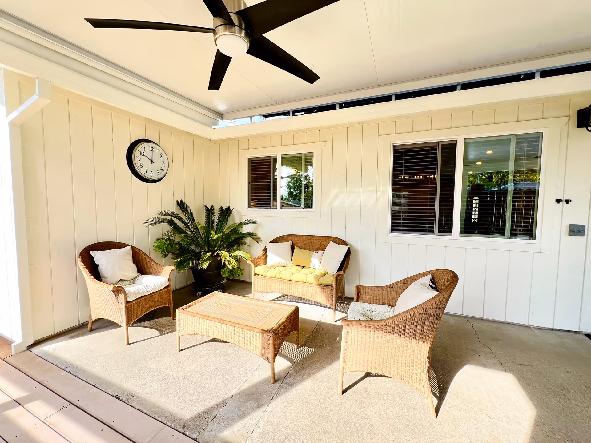 Covered outdoor patio area with wicker furniture including two armchairs, a loveseat with cushions, and a wicker coffee table. A large potted plant is placed in the corner, and a black ceiling fan is mounted on the white ceiling. A wall clock is visible on the cream-colored paneled wall, along with two windows with white frames and blinds.