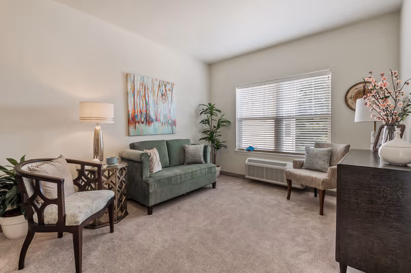 Bright furnished living room with a green loveseat, two chairs, a lamp, window blinds, and decorative plants.
