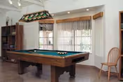 A communal game room with a green-felt pool table, stained-glass overhead light, bookshelves, vertical blinds, and a wooden chair.