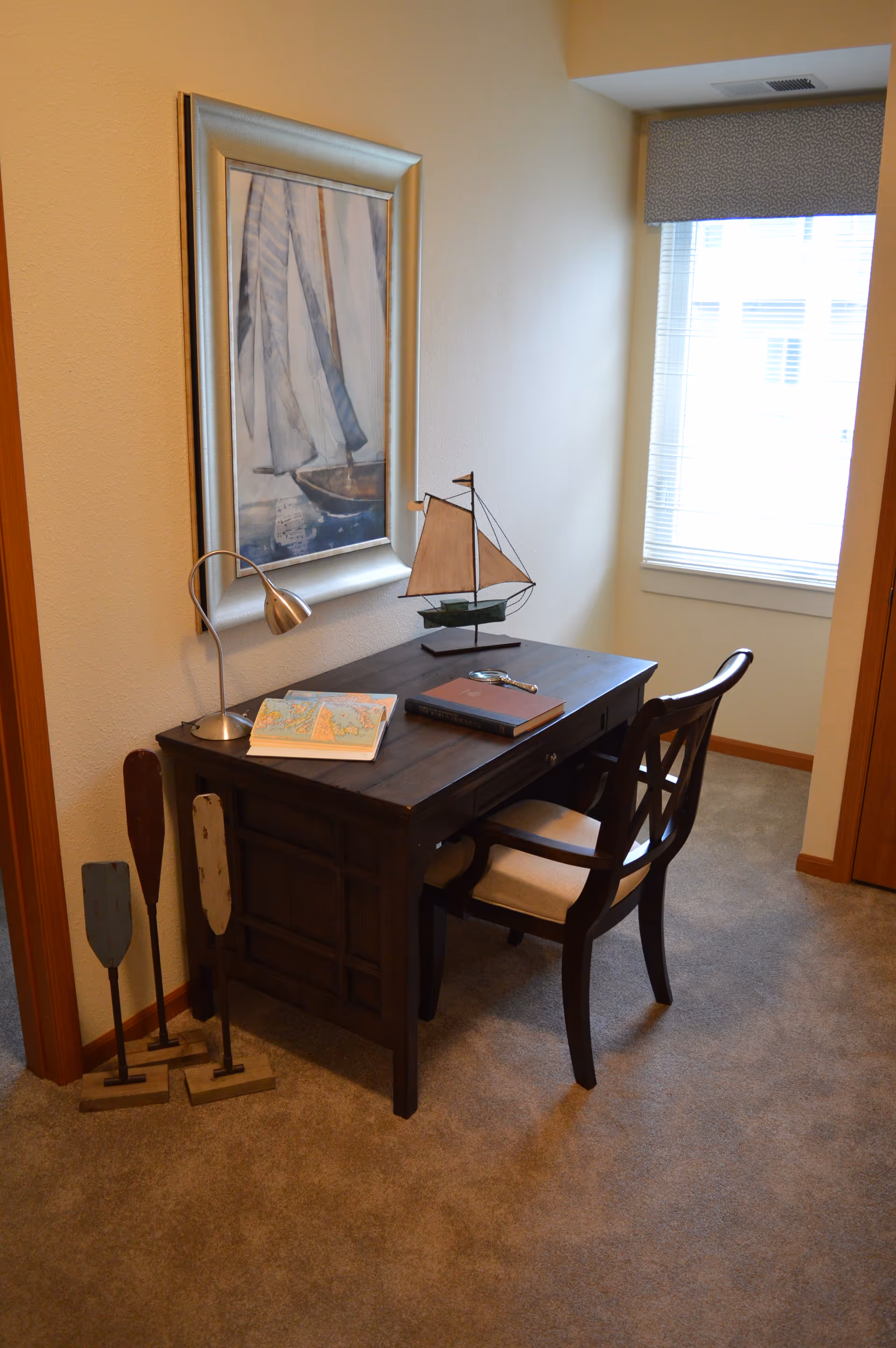 A small study area with a dark wooden desk and matching chair with a cushioned seat. On the desk are a desk lamp, an open book, a closed book, and a decorative model sailboat. Behind the desk is a framed painting of a sailboat. To the left of the desk are three decorative wooden oars standing upright. A window with blinds and a valance is on the right wall, letting in natural light. The room has beige walls and carpeted flooring.