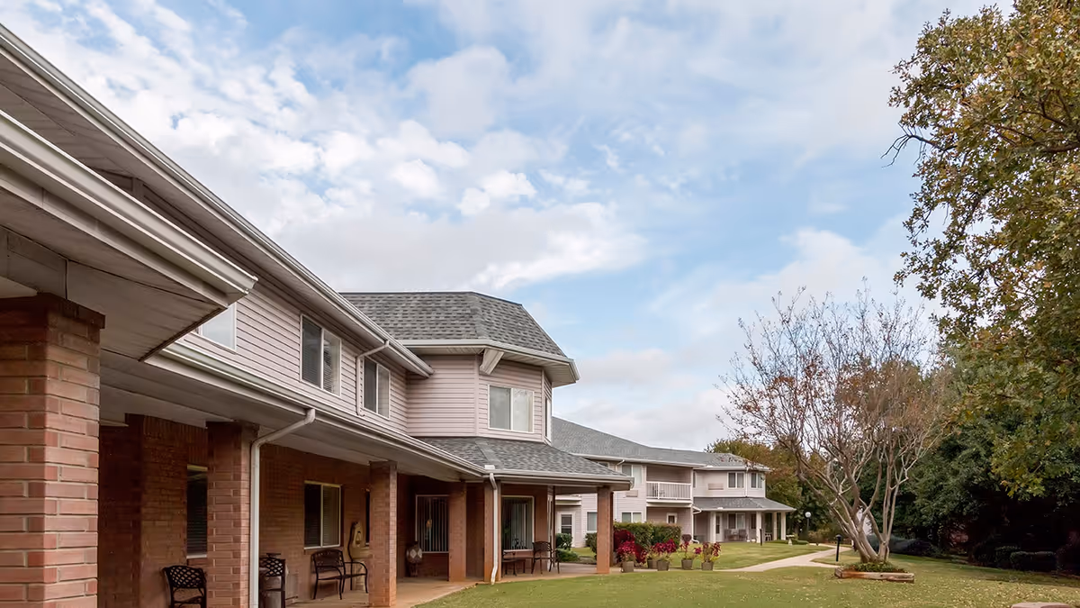 Exterior view of a senior living facility with a covered porch supported by brick columns, several chairs placed along the porch, a well-maintained lawn, potted plants, a tree with sparse leaves, and a partly cloudy sky.