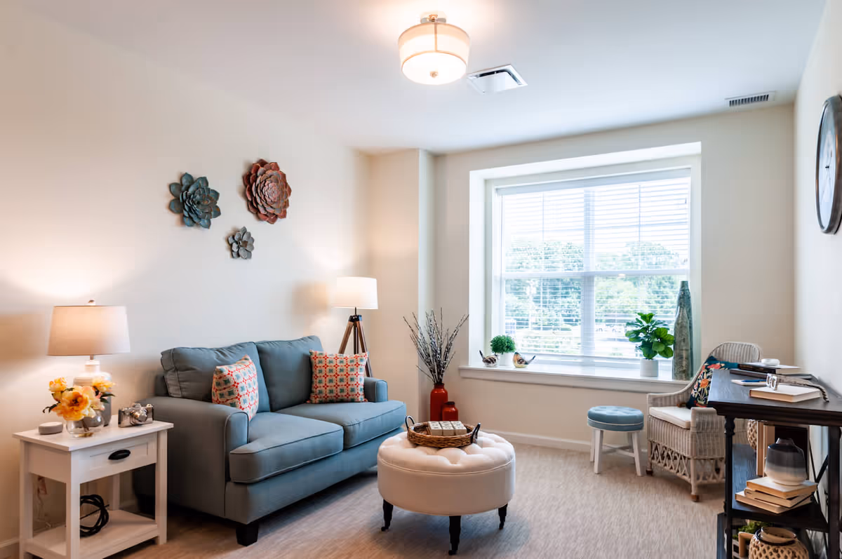 Bright, cozy living room with a blue loveseat, round ottoman, side tables, lamps, decorative wall art, and a large window seat.