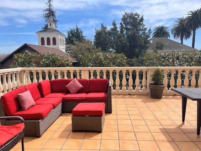 Outdoor patio area with terracotta tiled floor, featuring a brown wicker sectional sofa with red cushions and pillows, a matching ottoman, a wicker chair with a red cushion, a rectangular table, and a potted plant. In the background, there are trees, a building with a cupola, and a partly cloudy sky.