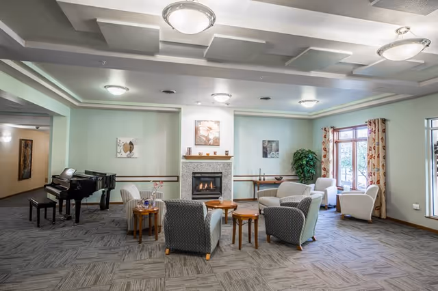A spacious and well-lit common area with a fireplace in the center, surrounded by comfortable armchairs and small wooden tables. There is a black grand piano on the left side and a large window with patterned curtains on the right, allowing natural light to enter. The walls are painted light green and decorated with framed artwork, and a potted plant is placed near the window.
