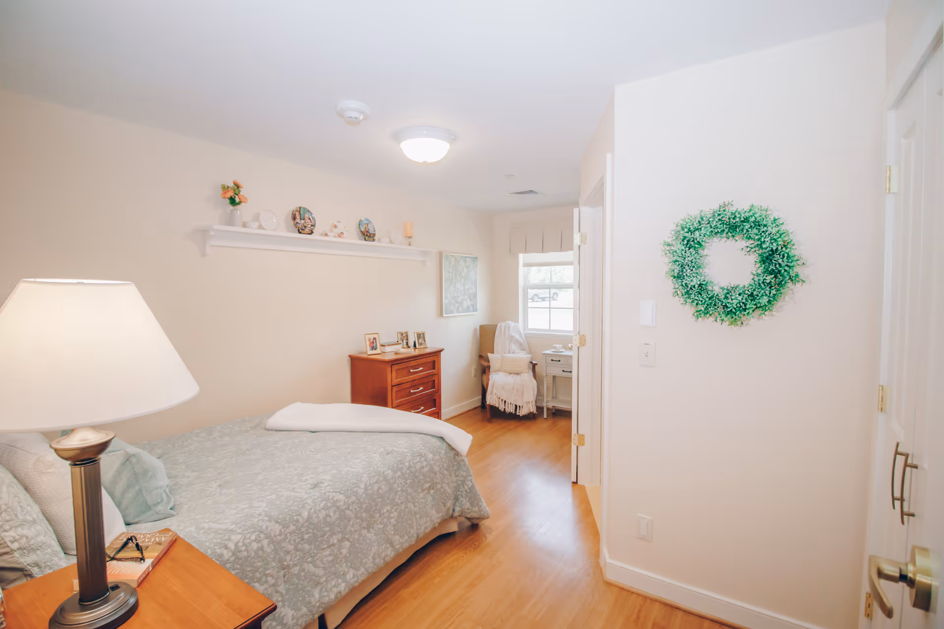 A cozy bedroom in a senior living facility with a bed covered in a light blue patterned comforter, a wooden nightstand with a lamp and a book, a wooden dresser with framed photos, a chair with a throw blanket near a window, and a green wreath hanging on the wall.
