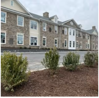 Front exterior of a two-story senior living building with a stone facade, multiple windows, and landscaped shrubs in the foreground.