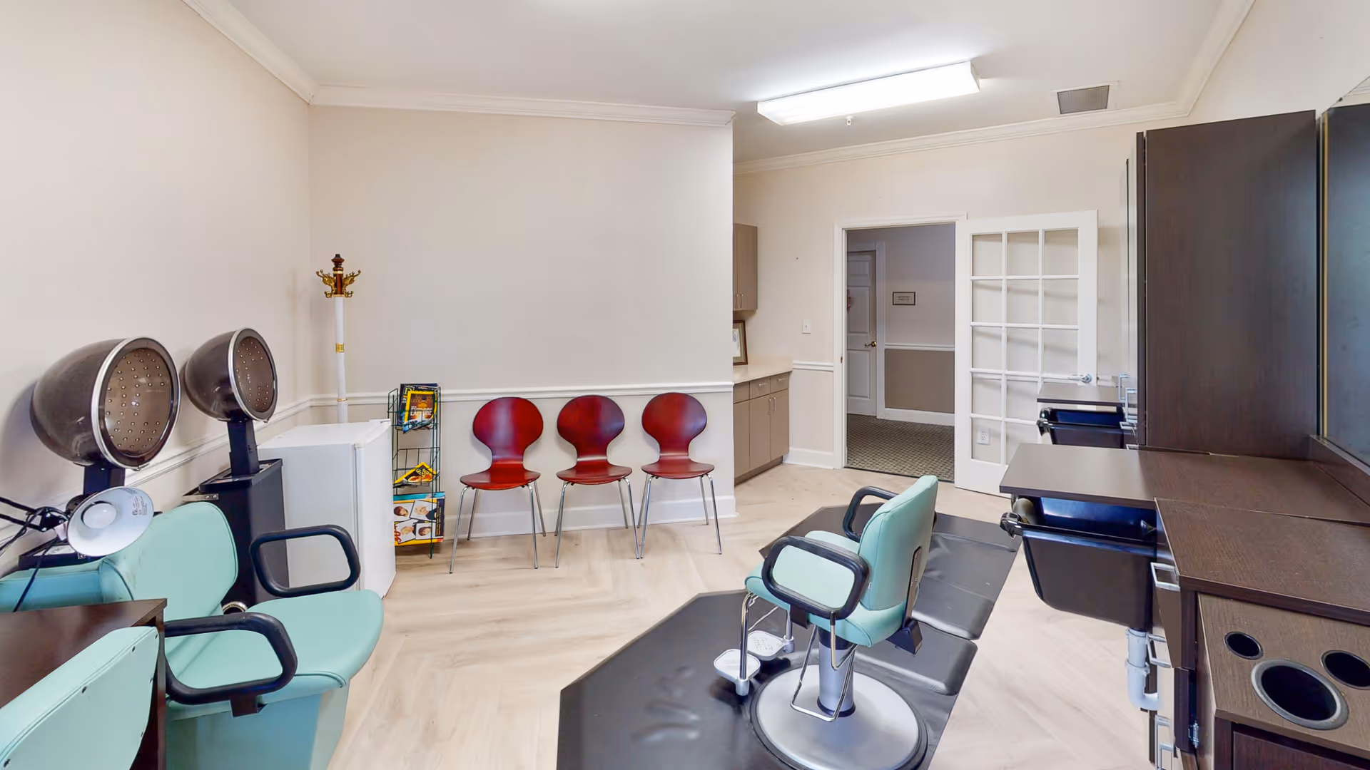 Interior of a salon area in a senior living facility with two green salon chairs, two hair dryers, a row of three red chairs against the wall, a coat rack, and a small white refrigerator. There is a door with glass panels leading to another room.