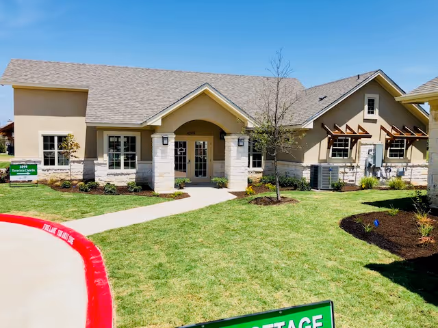 Exterior view of a single-story building with beige walls, a gray shingled roof, and a central entrance with double glass doors. The building is surrounded by a well-maintained lawn, small trees, and landscaped flower beds under a clear blue sky.