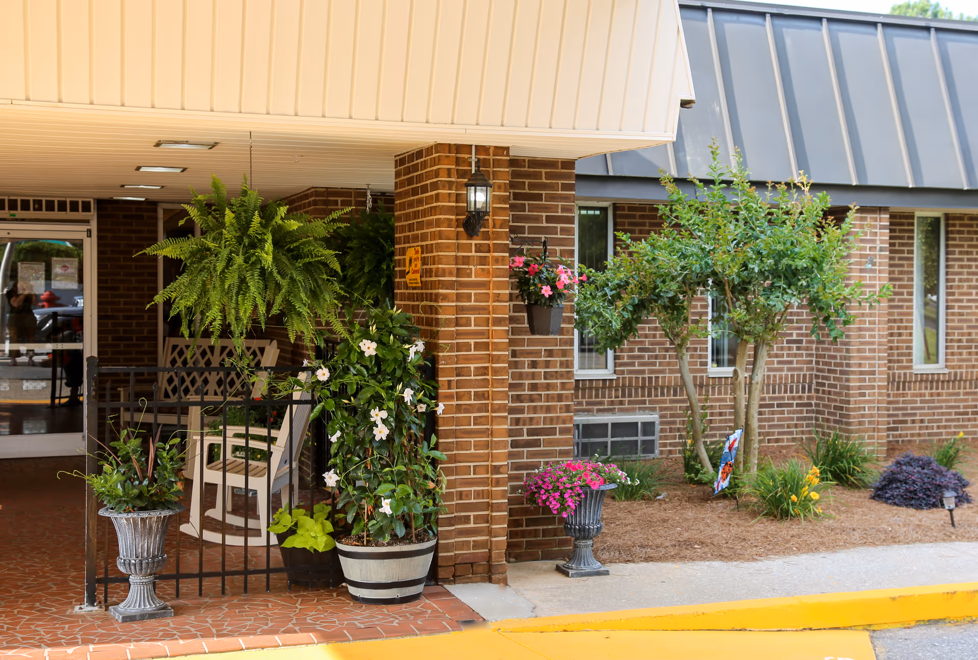 Covered brick facility entrance with potted flowers, hanging ferns, a rocking chair, and a small landscaped area.