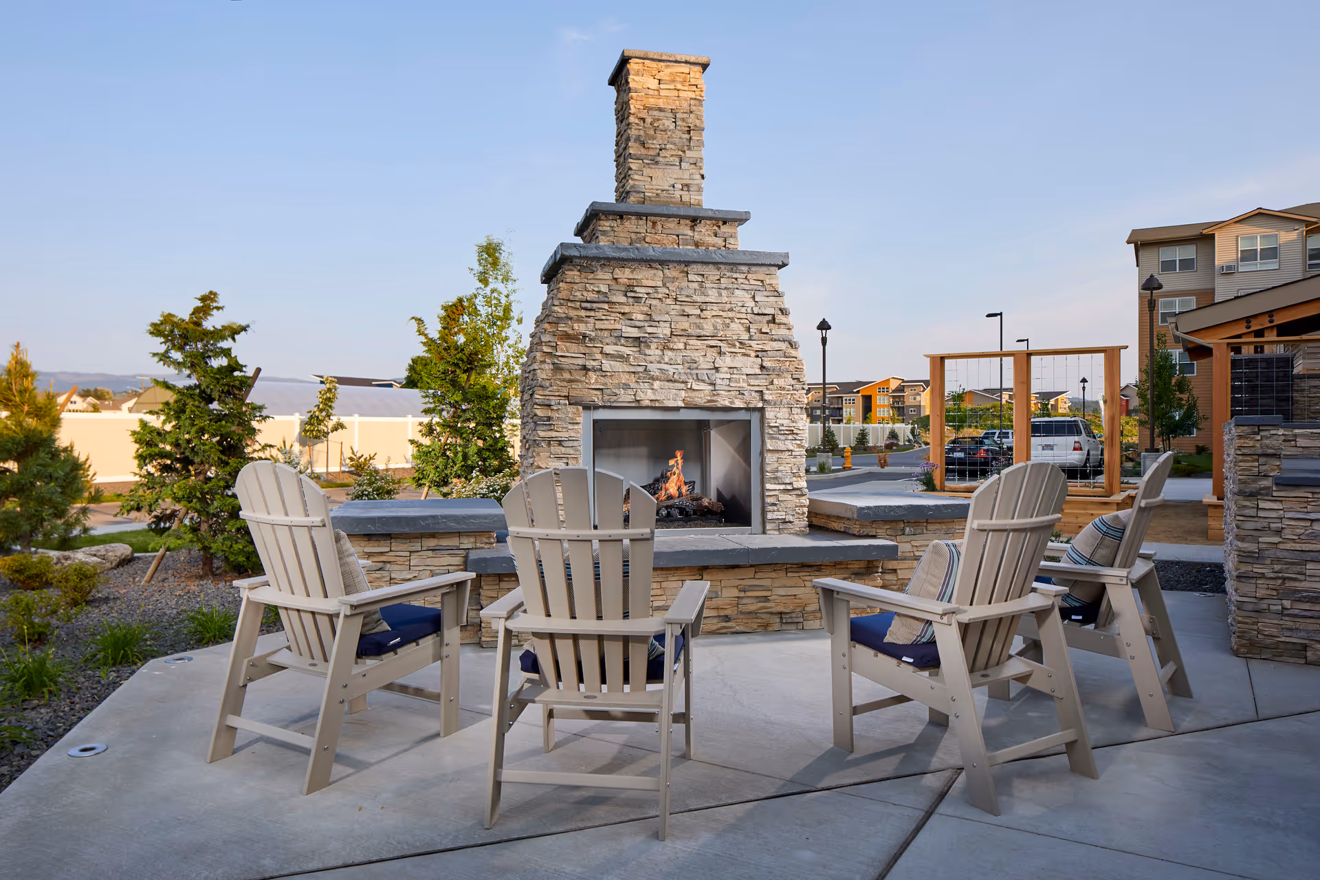 Outdoor seating area with four beige Adirondack chairs with cushions arranged around a stone fireplace with a fire burning. The area is paved with concrete and surrounded by landscaping with trees and shrubs. In the background, there are buildings and parked cars under a clear sky.