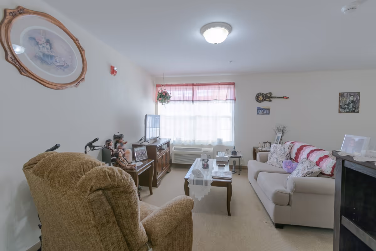 A cozy living room in a senior living facility with a beige recliner chair and a beige sofa adorned with patterned pillows and a pink and white crocheted blanket. A wooden coffee table with a lace runner sits in the center, holding tissues, a remote, and other small items. A TV is placed on a wooden cabinet against the wall, with decorative dolls and framed photos nearby. The room has a large window with sheer white curtains and a pink valance, and the walls are decorated with framed pictures and a guitar-shaped wall clock.