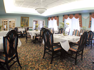 Dining room with several round tables set with white tablecloths and wooden chairs, large windows with valances, and floral carpet.