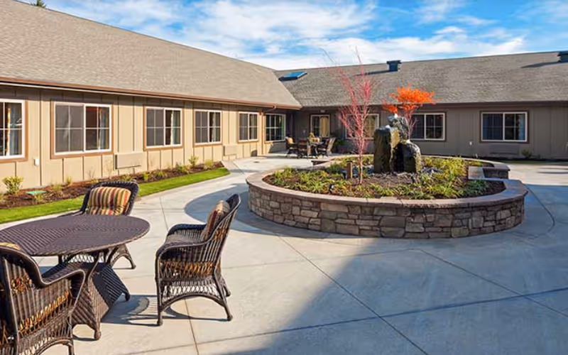 Sunny courtyard patio with wicker tables and chairs around a circular stone planter and single-story building.