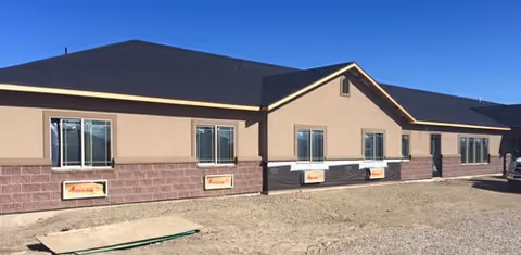 Exterior view of a single-story building with beige and brown walls, multiple windows, and a dark roof under a clear blue sky. The ground in front is bare dirt with some construction materials.