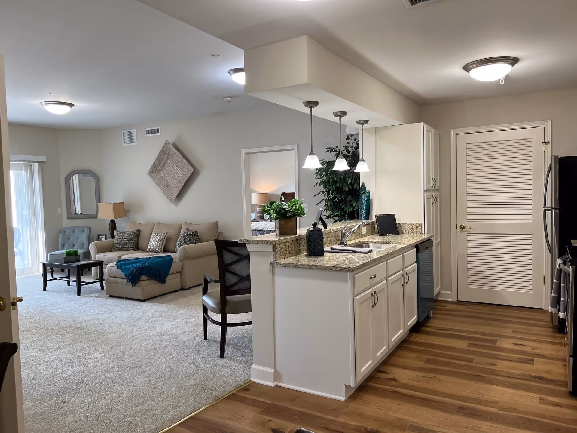 Interior view of a senior living facility apartment showing a kitchen with white cabinets, granite countertops, and pendant lights. Adjacent to the kitchen is a living room area with a beige sofa, a blue throw blanket, a coffee table, a side table with a lamp, and a blue armchair. The floor transitions from wood in the kitchen to carpet in the living room. A doorway leads to a bedroom visible in the background.
