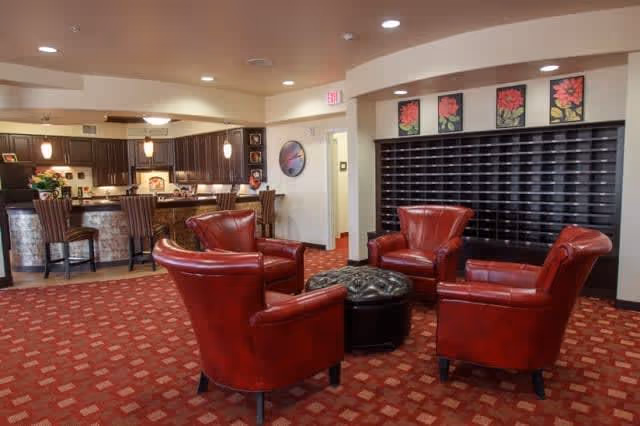A cozy common area with four red leather armchairs arranged around a black tufted ottoman on a patterned red carpet. In the background, there is a kitchen area with a counter and bar stools, dark wooden cabinets, and pendant lights. On the wall behind the chairs, there are four framed floral artworks above a large set of mailboxes or cubbies.
