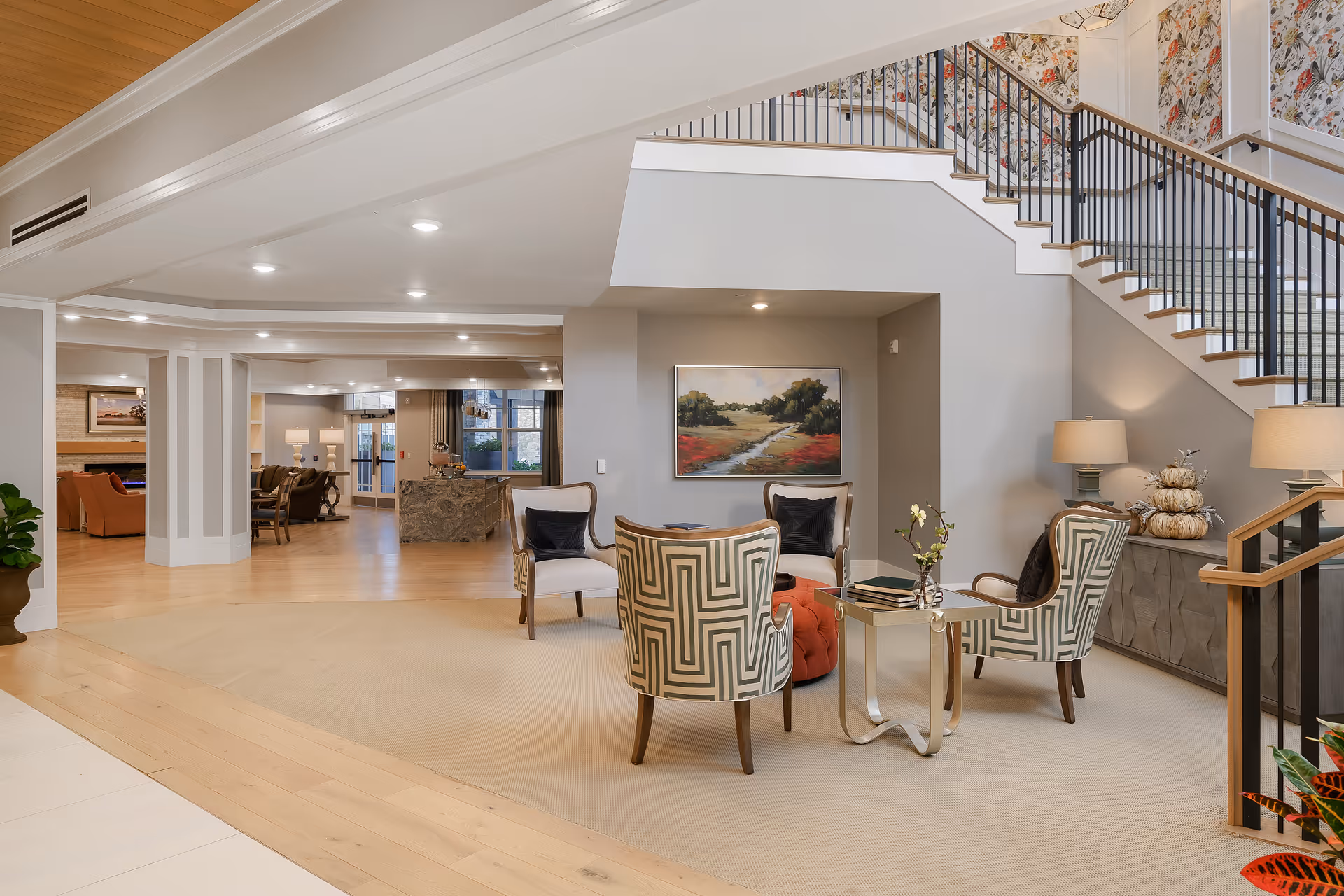 Spacious and well-lit senior living facility common area with a seating arrangement of four patterned armchairs around a small table on a beige carpet. The area features a staircase with black railings, a sideboard with lamps and decorative pumpkins, and a landscape painting on the wall. The background shows more seating areas, a reception desk, and large windows.
