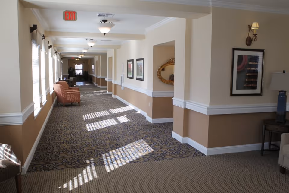 A long, well-lit hallway in a senior living facility with patterned carpet, beige walls with white trim, several framed pictures on the walls, and a few chairs placed along the corridor near windows letting in natural light.