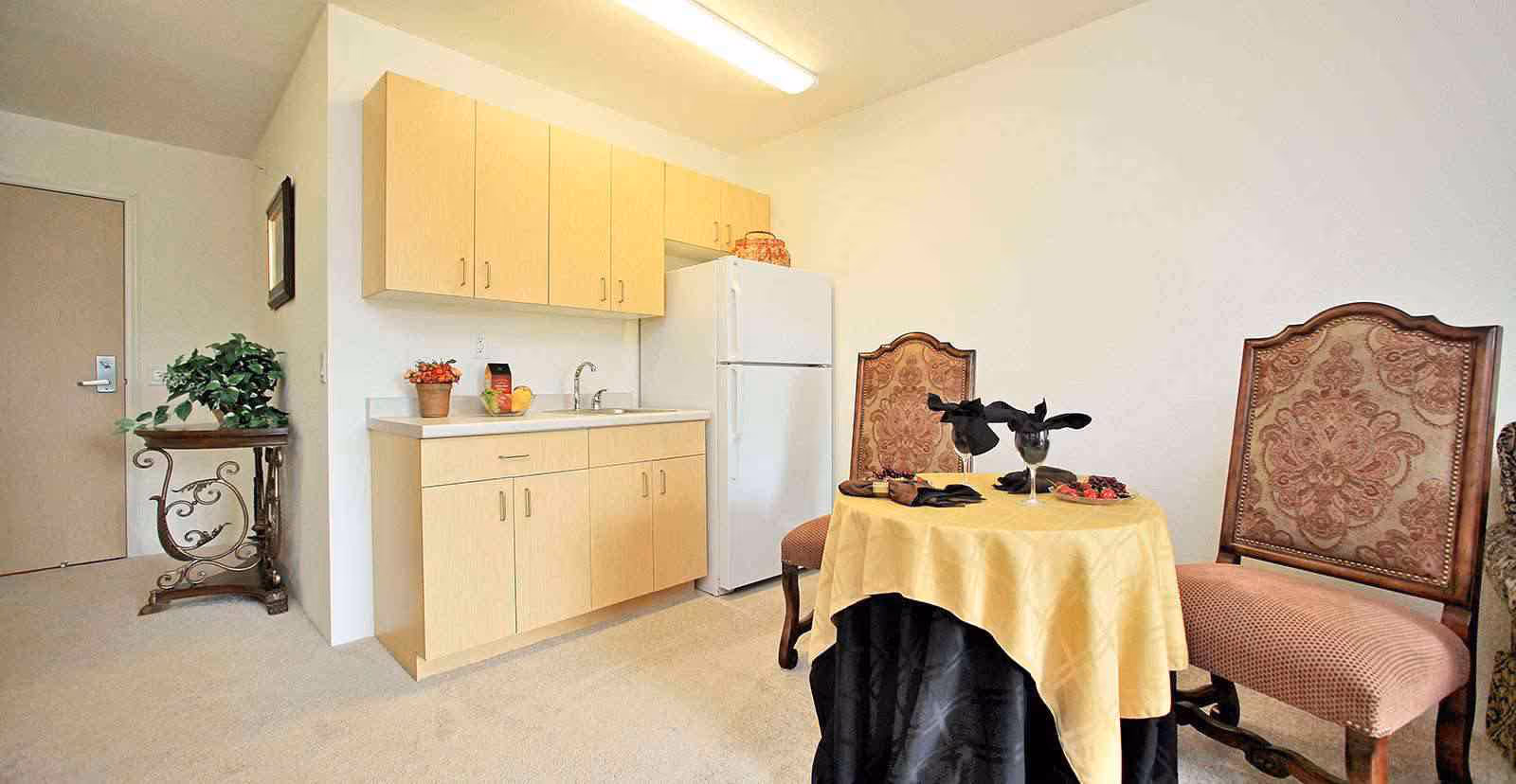 Small apartment-style kitchenette with light wood cabinets and a refrigerator beside a small round table set with two ornate chairs.