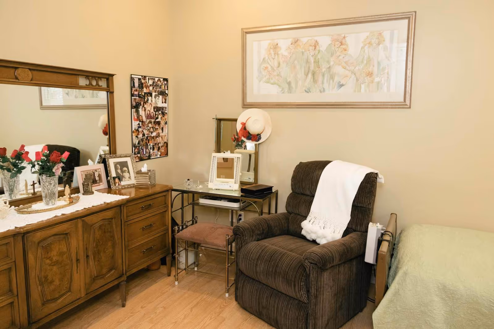 A cozy room with a brown upholstered armchair draped with a white throw blanket, a wooden dresser with a large mirror, framed photos, and a vase of red roses. Next to the armchair is a small glass table with a cushioned stool underneath, a mirror, and a decorative hat hanging on the wall. A bed with a green quilt is partially visible on the right side. The walls are beige, and a framed artwork hangs above the armchair.