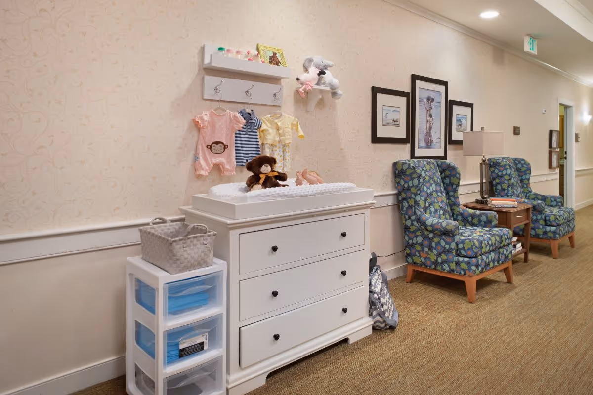 A hallway area in a senior living facility featuring a white changing table with baby clothes and stuffed animals on top, a small plastic drawer unit with a basket, two patterned armchairs with a small wooden table and lamp between them, and framed pictures on the wall.