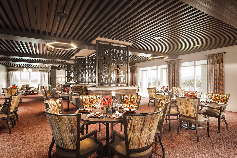 Bright, upscale dining room with multiple round tables set with plates and floral centerpieces, patterned chairs, decorative screens, and large windows.