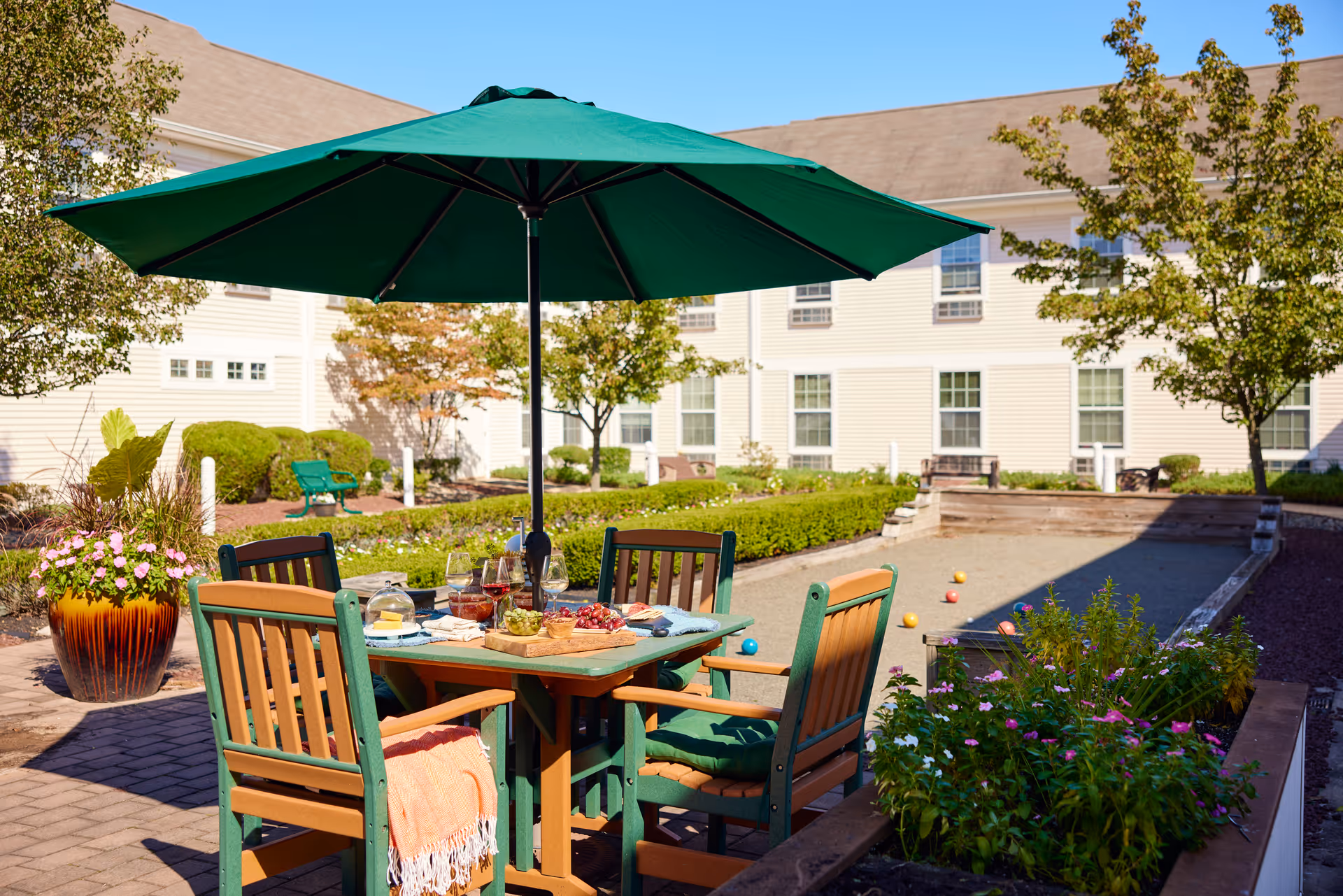 Outdoor patio area with a table and four chairs under a large green umbrella. The table has snacks and drinks on it. In the background, there is a bocce ball court with several bocce balls scattered on it, surrounded by greenery and a beige building with multiple windows.