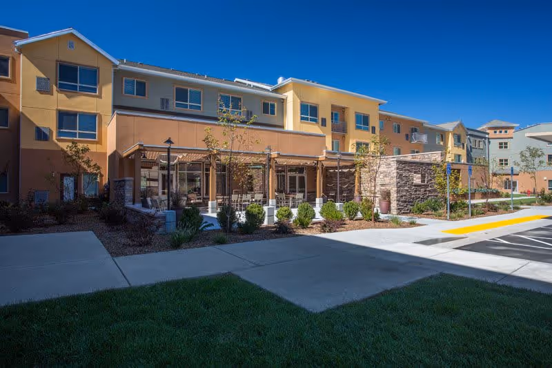 Exterior view of a multi-story senior living facility building under a clear blue sky, featuring a landscaped garden area with shrubs and trees, a covered patio with seating, and a paved walkway and parking area in the foreground.