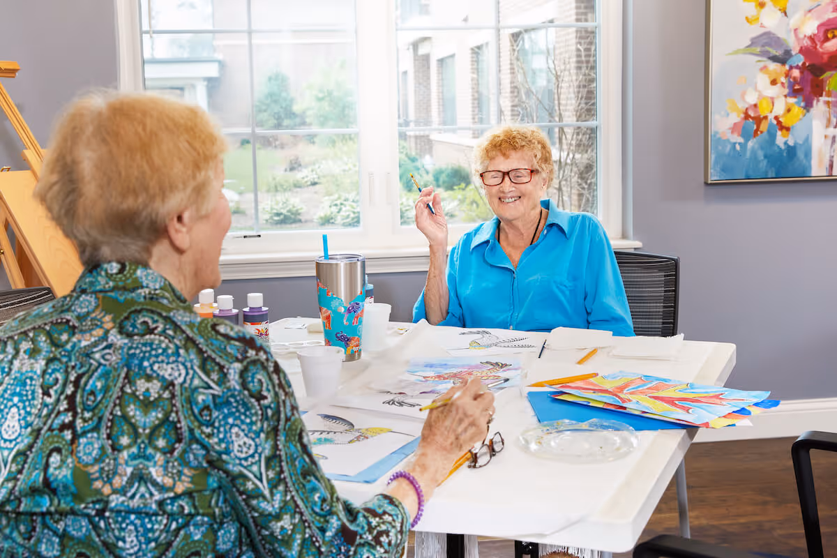 Two elderly women sitting at a table in a bright room with large windows, engaging in painting and drawing activities. One woman is wearing a blue shirt and glasses, smiling while holding a paintbrush, and the other woman is seen from behind wearing a patterned green shirt. The table is covered with art supplies, colorful paintings, and cups.