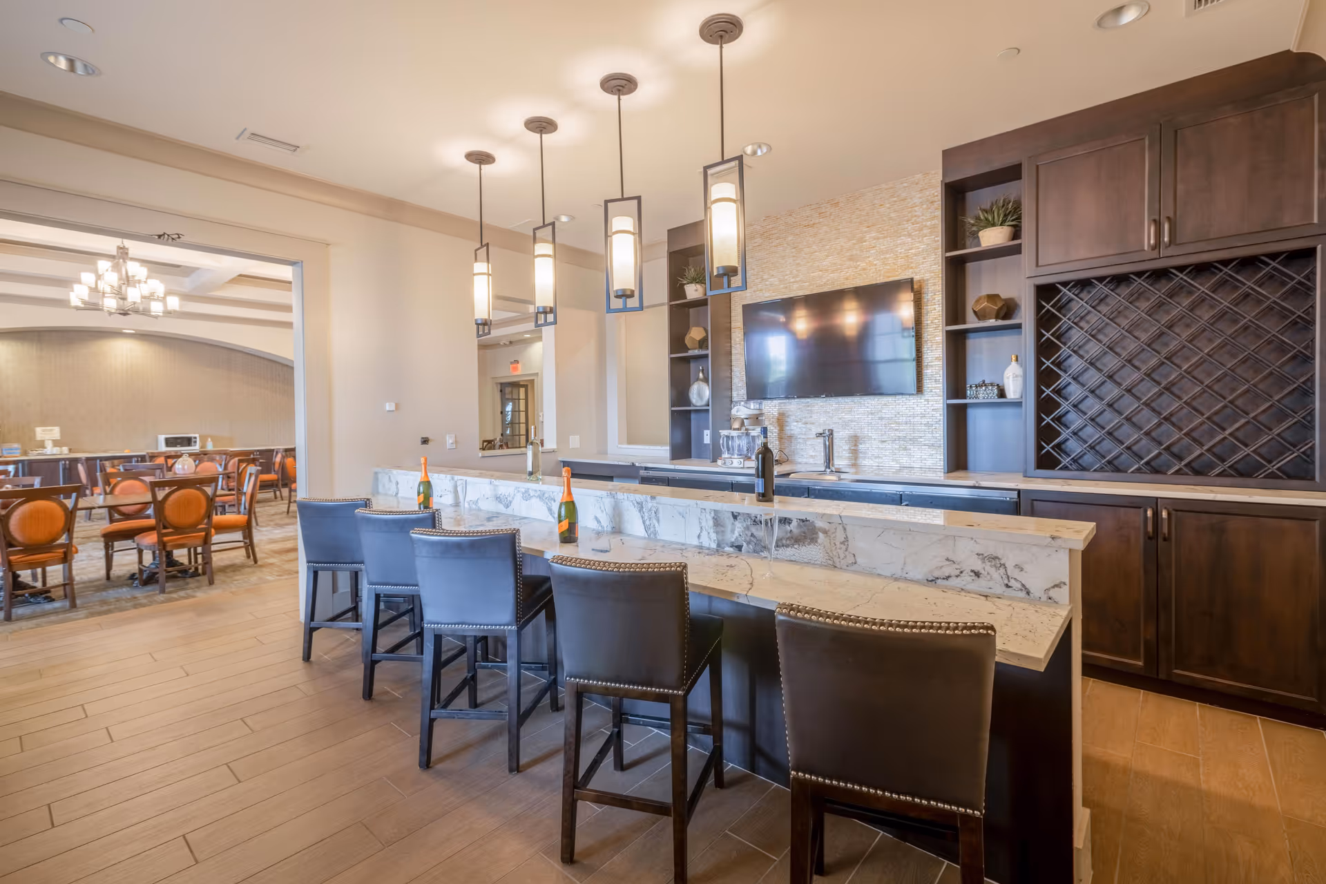Interior view of a senior living facility's bar area with a marble countertop and five high chairs. The bar has pendant lights hanging from the ceiling, a mounted flat-screen TV, and dark wooden cabinets with shelves. In the background, there is a dining area with round tables and chairs, and a chandelier hanging from the ceiling.