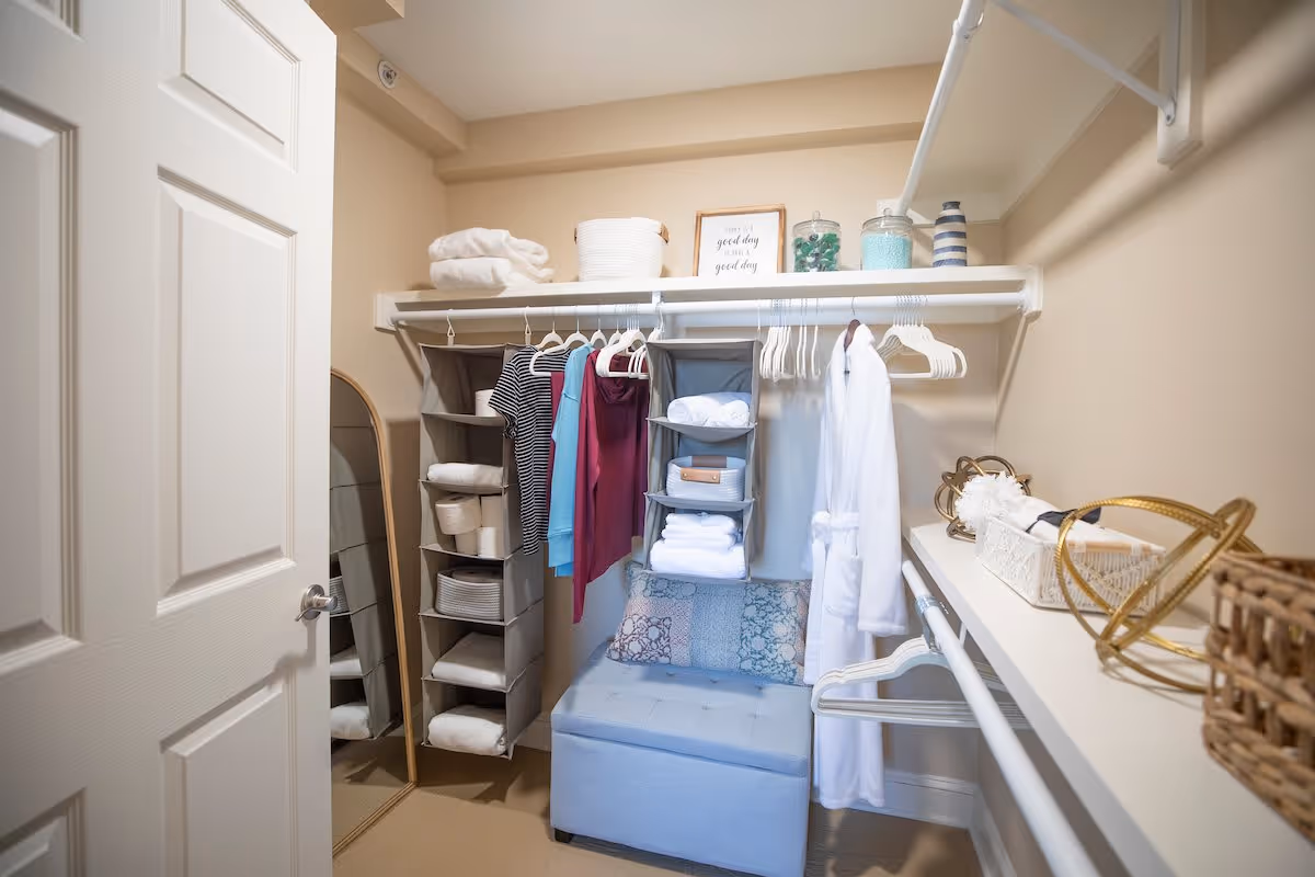 A neatly organized walk-in closet with hanging clothes including shirts and a white robe, shelves holding folded towels and decorative jars, a full-length mirror leaning against the wall, and a cushioned blue storage bench with patterned pillows.