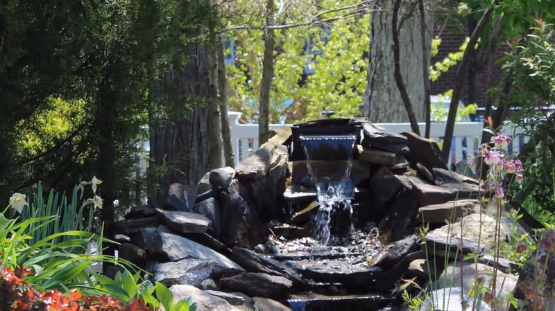 A small cascading water feature made of rocks surrounded by green plants and flowers in an outdoor garden setting with trees and a white fence in the background.
