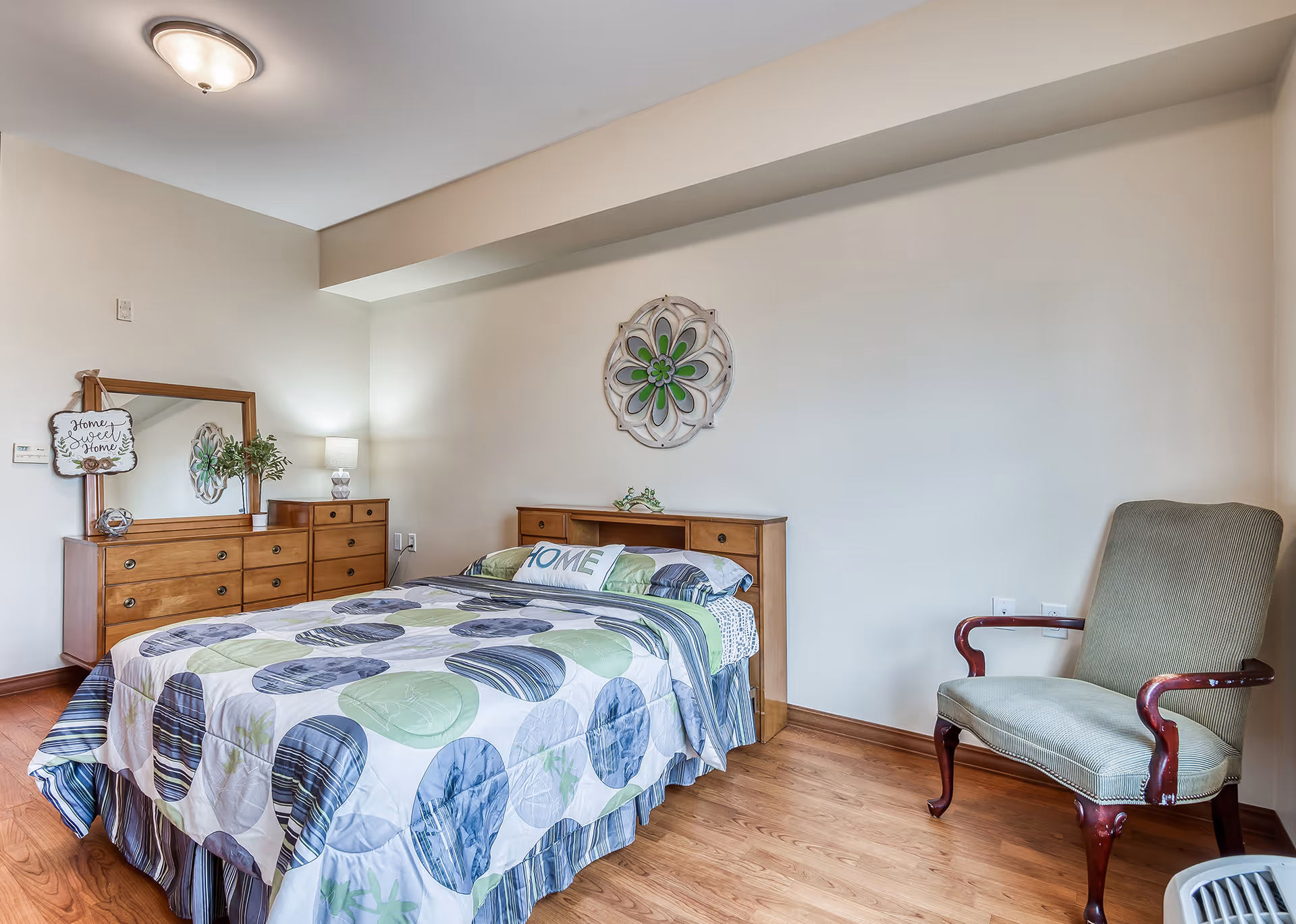 A cozy bedroom with a wooden bed featuring a colorful circular patterned bedspread and a pillow with the word 'HOME'. There is a wooden dresser with a mirror and a lamp on top, decorated with a small plant and a sign that reads 'Home Sweet Home'. A cushioned armchair with wooden armrests is placed near the bed. The room has light-colored walls and wooden flooring.