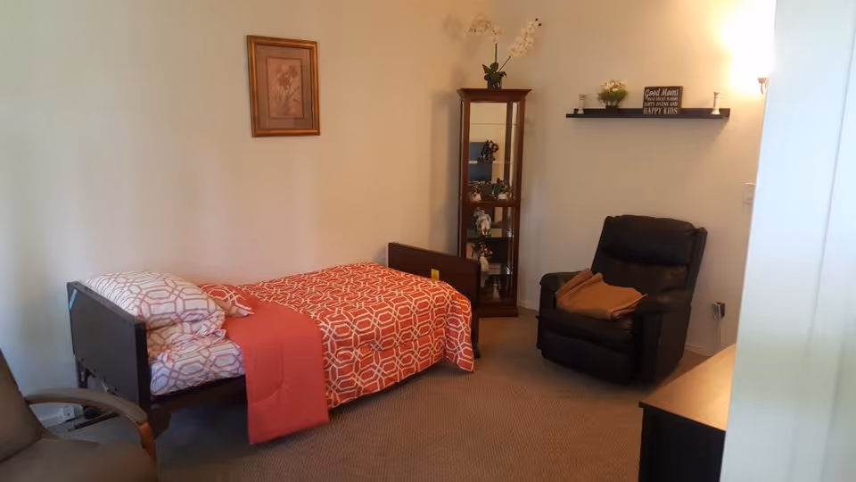 A cozy bedroom in a senior living facility with a single bed covered in a red and white patterned blanket and pillows, a black recliner chair with a brown blanket, a wooden display cabinet with decorative items, a wall shelf with small plants and a sign, and a framed floral picture on the wall.