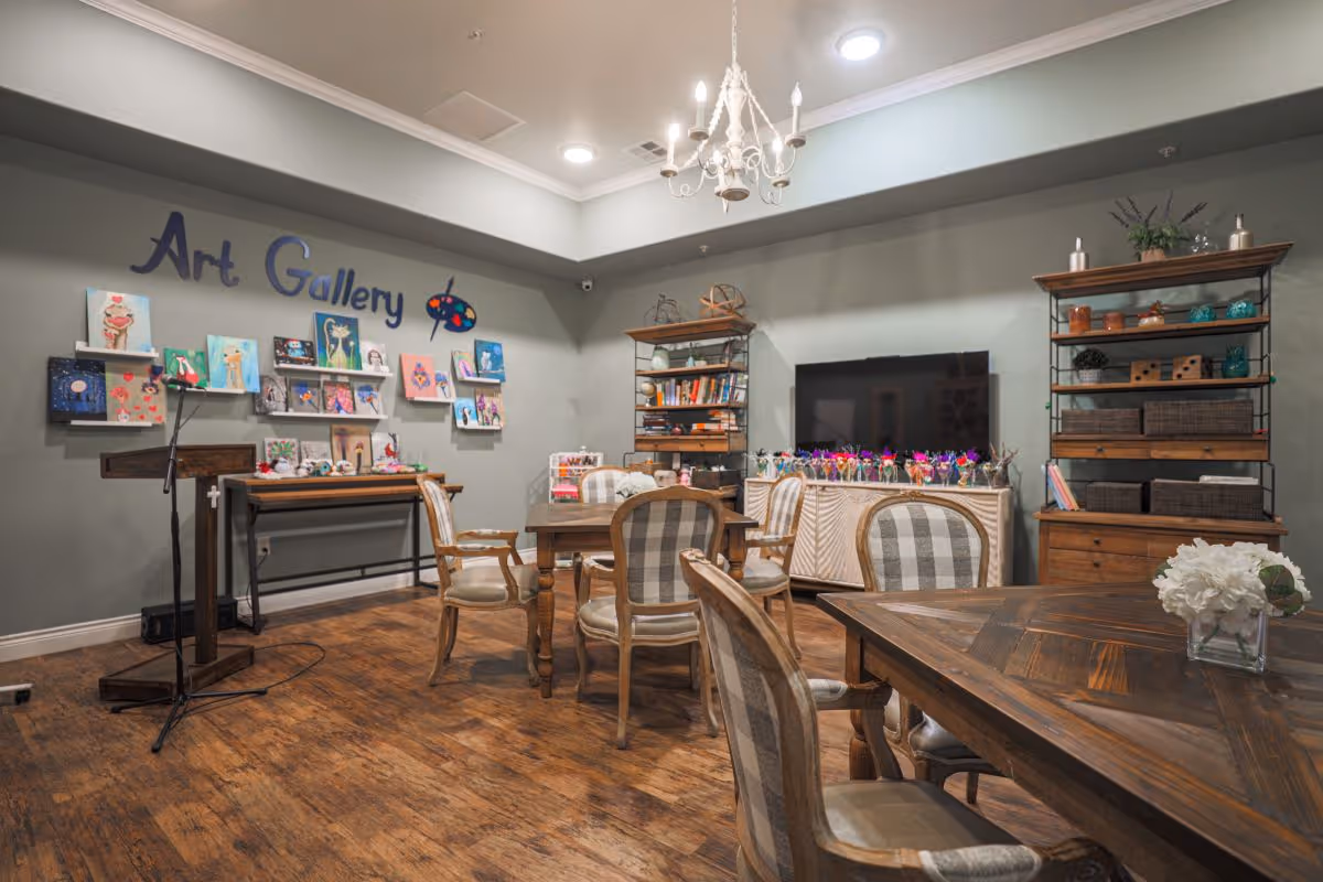 A cozy interior room labeled as an Art Gallery with several paintings displayed on the wall. The room features wooden floors, multiple wooden tables and chairs with cushioned seats, bookshelves with decorative items, a large flat-screen TV, and a chandelier hanging from the ceiling.