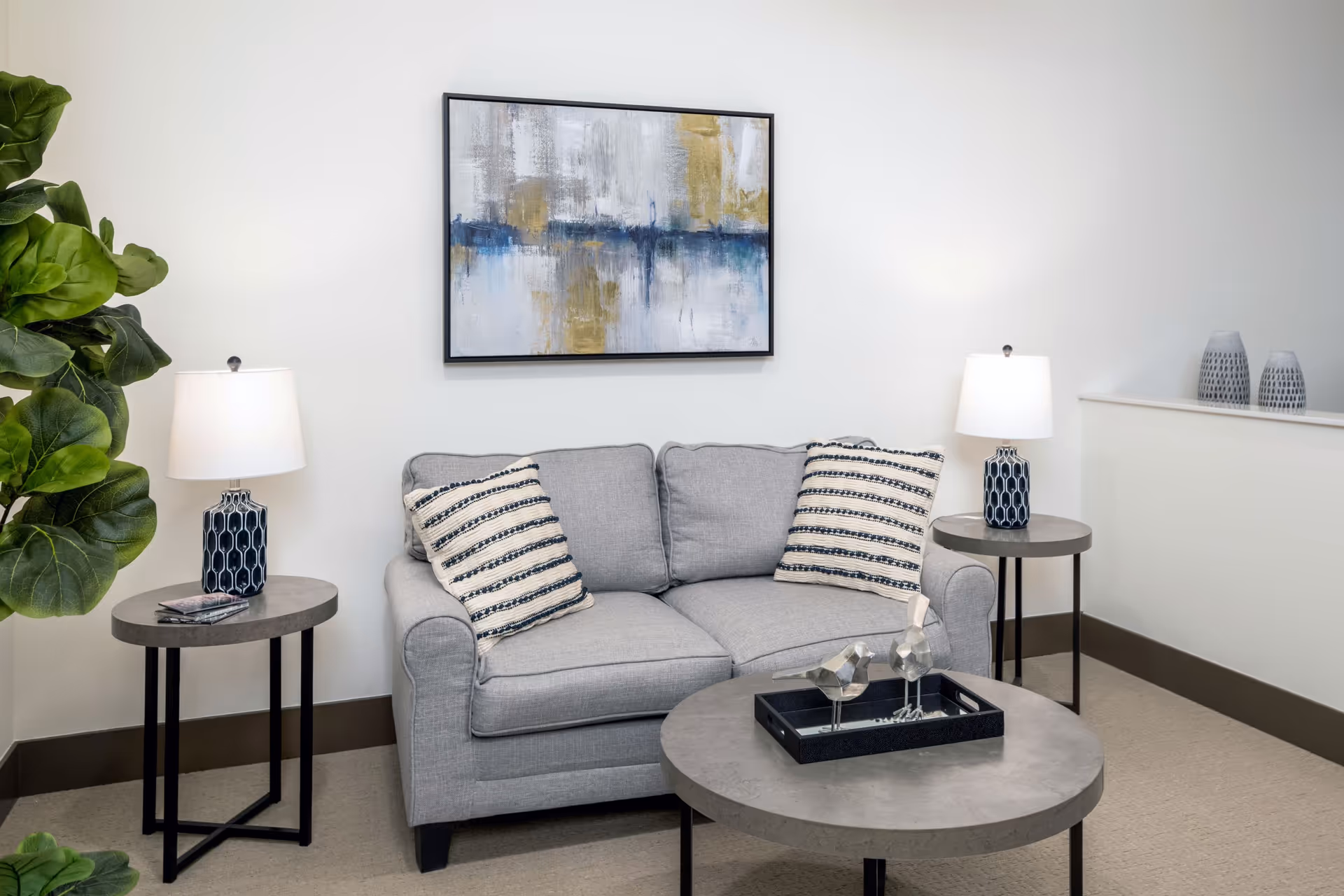 A cozy living room area with a light gray sofa adorned with two striped cushions. On either side of the sofa are round side tables with matching blue and white patterned lamps. A round coffee table in front of the sofa holds a black tray with two decorative silver bird figurines. A large abstract painting with blue, gray, and gold tones hangs on the white wall behind the sofa. A green leafy plant is visible on the left side of the image.