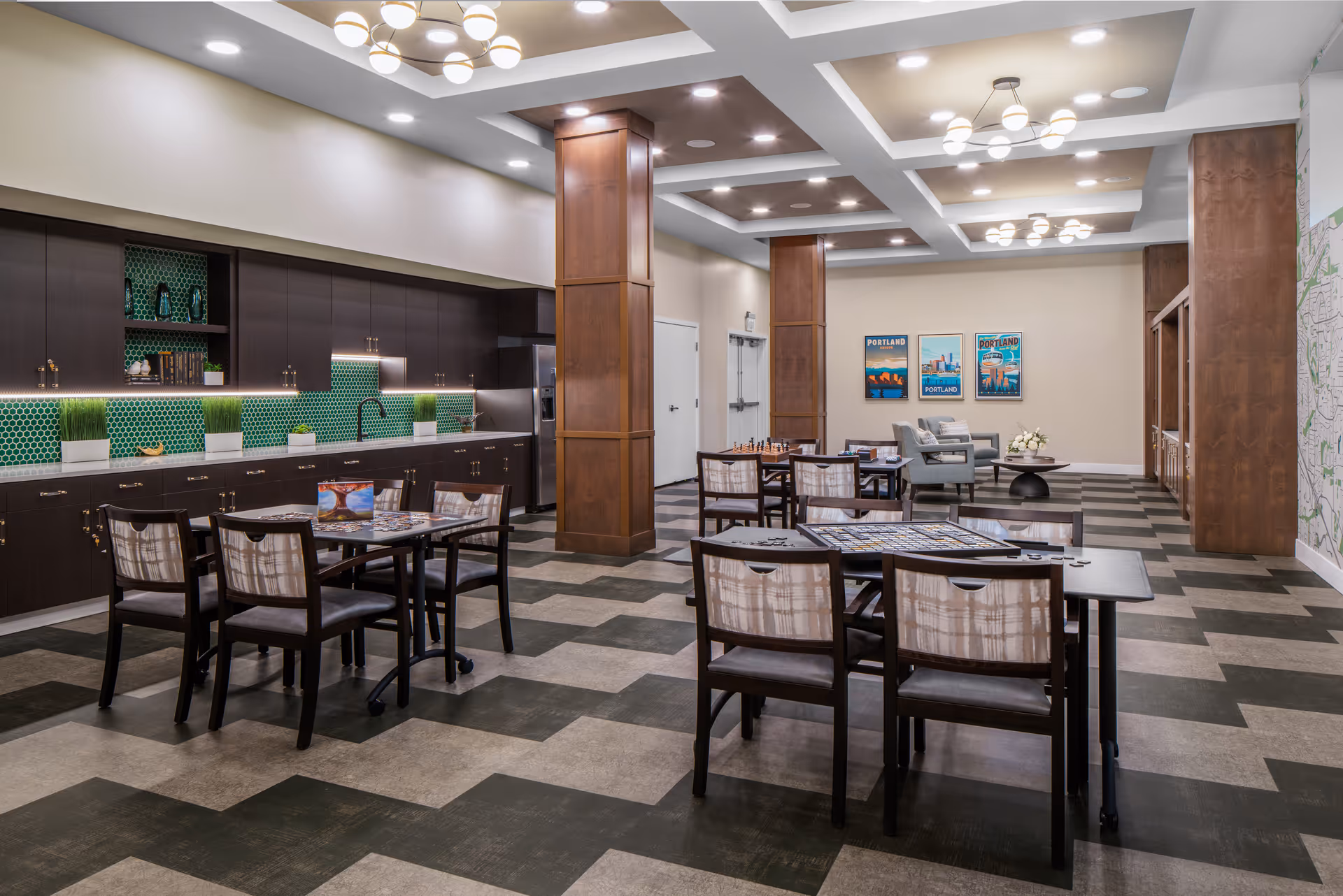 A spacious, well-lit common area in an assisted living facility featuring multiple tables and chairs arranged for games and social activities. The room has a modern design with dark wood cabinetry along one wall, green hexagonal tile backsplash, and a kitchenette area. There are decorative wooden columns, ceiling lights, and framed artwork on the far wall. Comfortable seating and a coffee table are visible in the background.