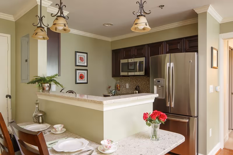 A modern kitchen area with light green walls, dark wood cabinets, and stainless steel appliances including a refrigerator and microwave. There is a countertop with a vase of red flowers and place settings with plates and teacups. Two pendant lights hang from the ceiling above the counter.