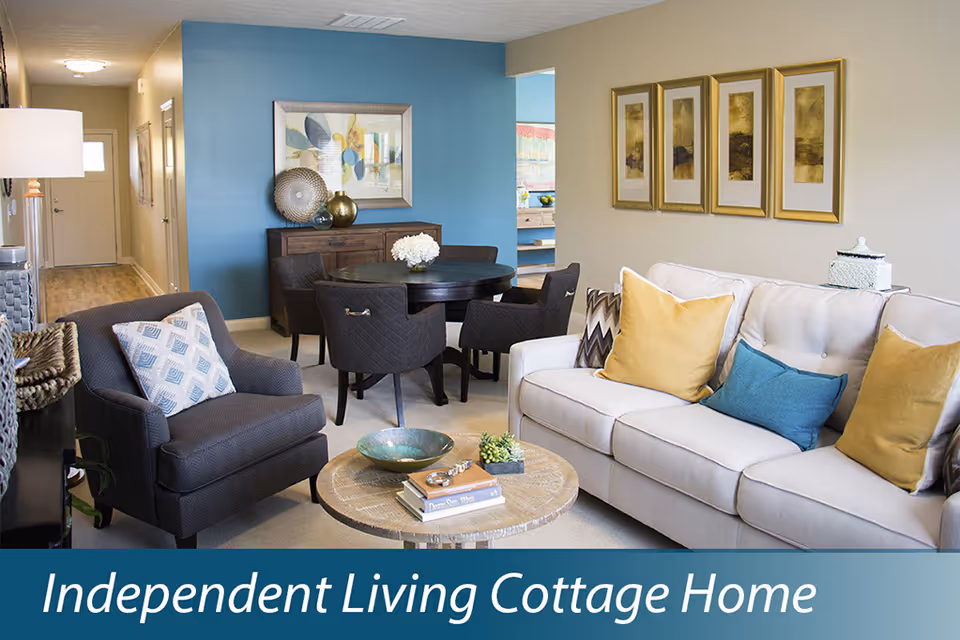 A cozy living room and dining area in an independent living cottage home. The living room features a light-colored sofa with yellow and blue pillows, a round wooden coffee table with decorative items, and a dark upholstered armchair with a patterned pillow. The dining area has a round dark wooden table with four matching chairs and a sideboard against a blue accent wall with framed artwork above it. The space is well-lit and decorated with neutral and warm tones.