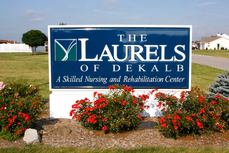 A large blue sign reading "The Laurels of DeKalb: A Skilled Nursing and Rehabilitation Center" stands on a white base with red flowers and lawn in front.