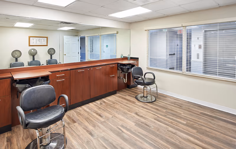 Interior view of a hair salon area in a senior living facility with wooden flooring, two black salon chairs, hair washing sinks, a long wooden cabinet with drawers, and a large mirror on the wall reflecting the room. There are windows with blinds and a door in the background.