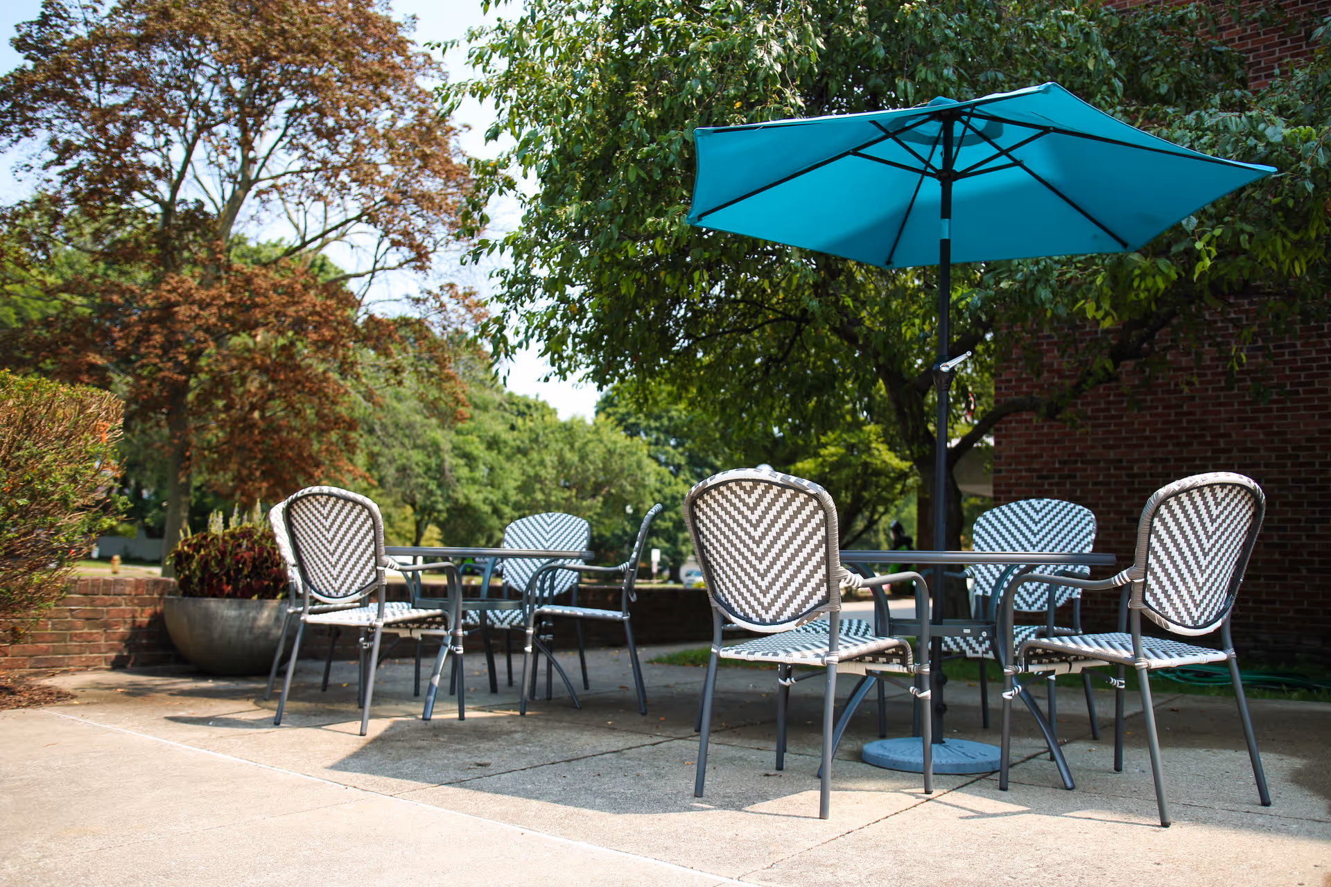 Outdoor patio area with a round glass table surrounded by six patterned chairs. A large blue umbrella provides shade. The setting is surrounded by trees and greenery with a brick building partially visible in the background.