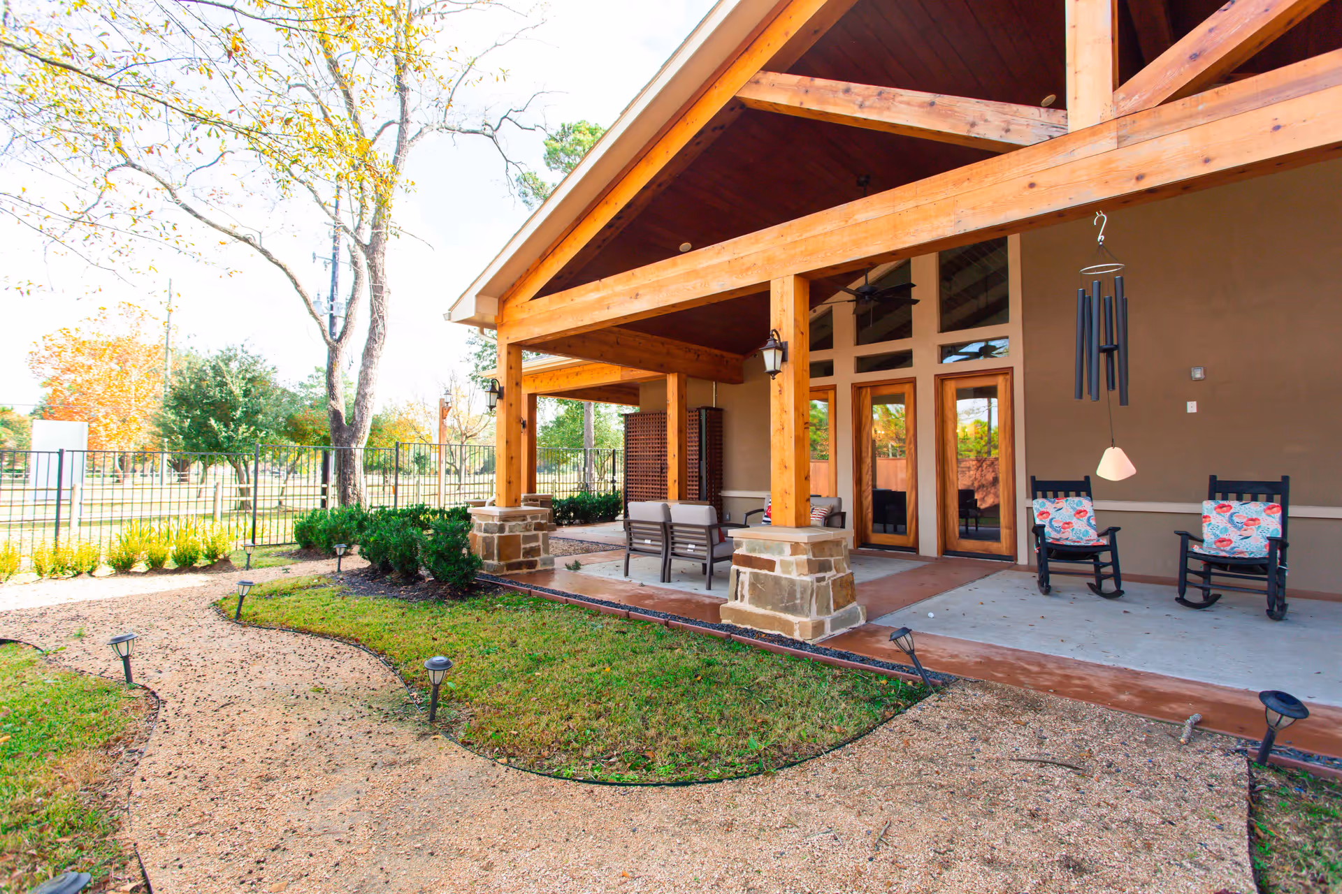 Covered exterior patio with wooden beams, seating and rocking chairs beside a landscaped gravel path and yard.