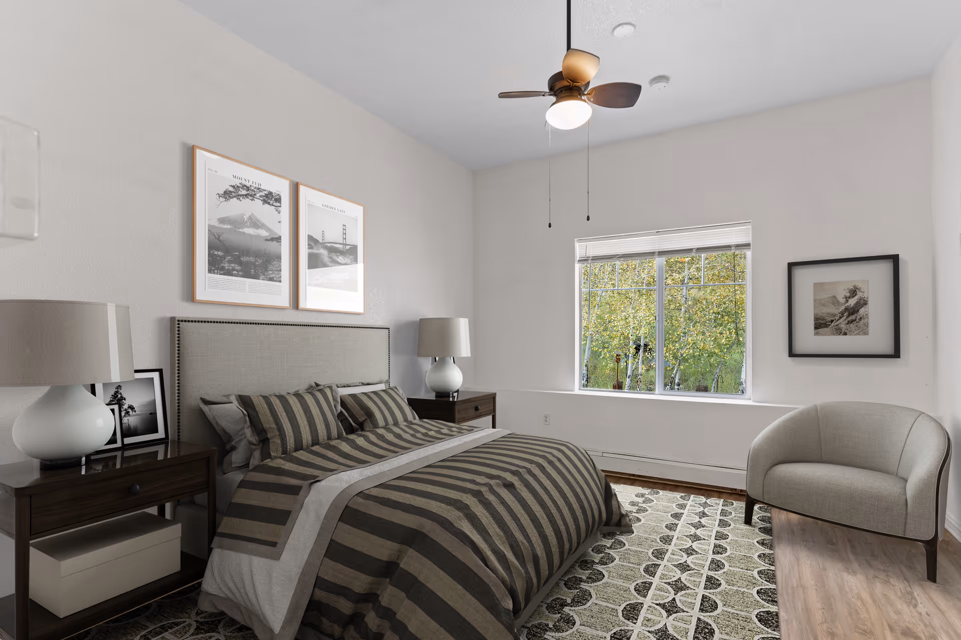A bedroom with a large bed featuring striped bedding and multiple pillows. There are two matching bedside tables with lamps on each side of the bed. Above the bed, two framed black and white pictures hang on the wall. A window with a view of green trees is on the right wall, and a cushioned armchair is positioned near the window. The room has a ceiling fan with a light and a patterned area rug on the wooden floor.