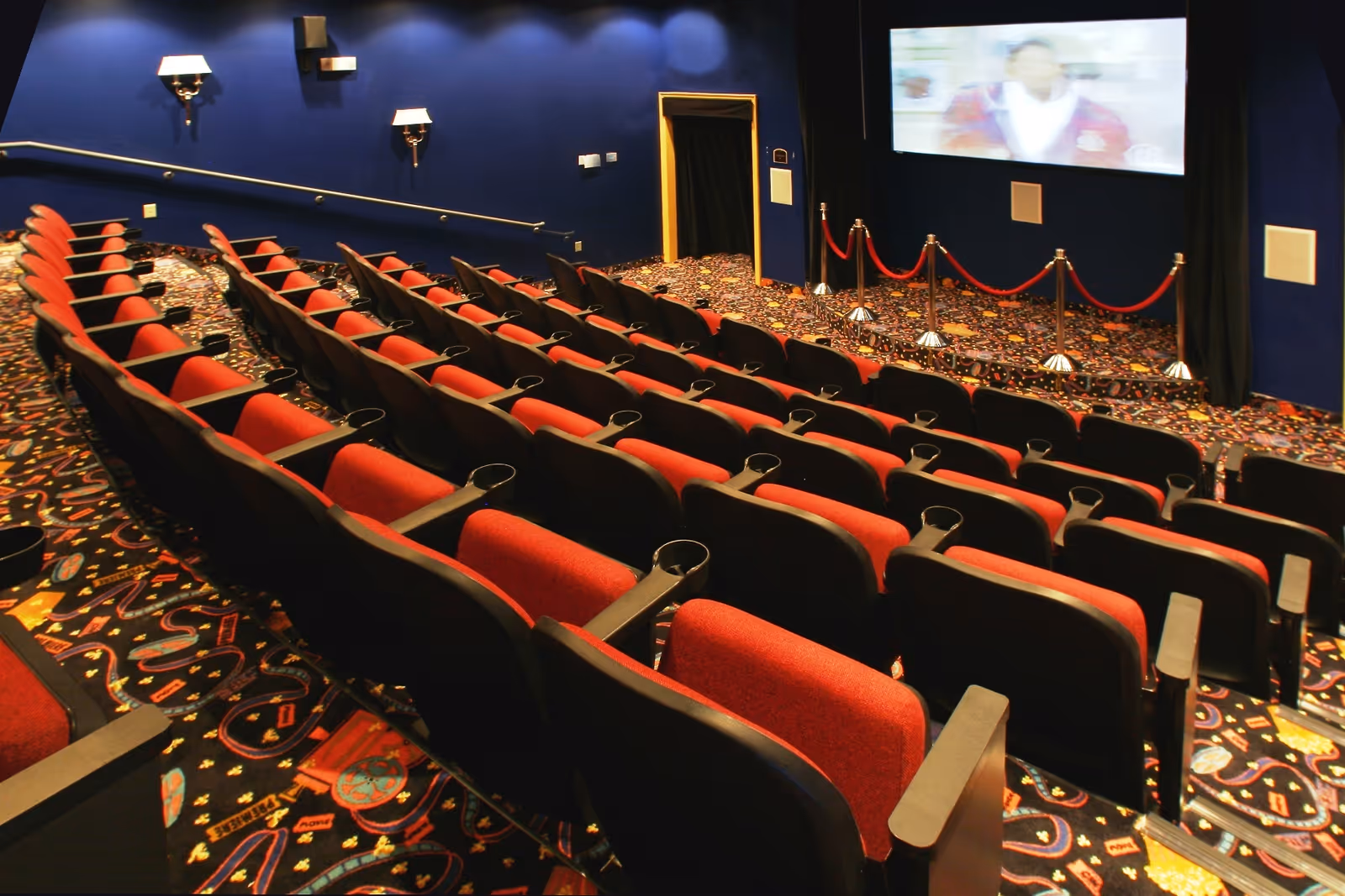 Interior view of a small movie theater with rows of red and black seats facing a large screen. The walls are dark blue with wall-mounted lights, and the floor is covered with a colorful patterned carpet. A red rope barrier is placed in front of the screen.