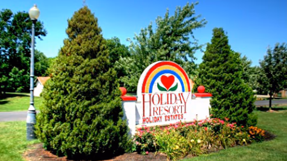 A colorful sign for Holiday Resort Holiday Estates surrounded by green trees and flowers, with a clear blue sky in the background and a street lamp on the left side.